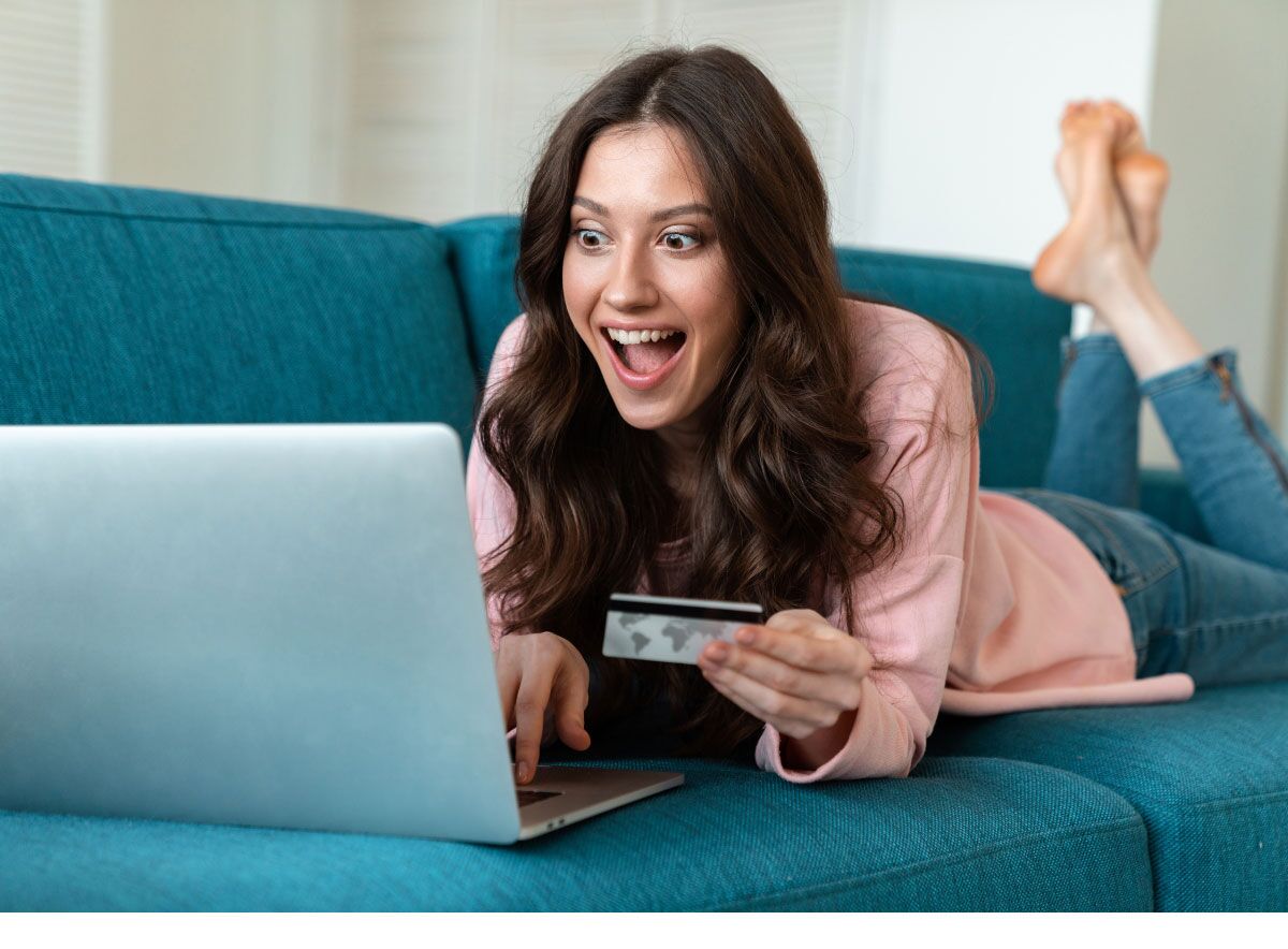 Woman laying on a couch holding a credit card and looking at a laptop is excited about a refund