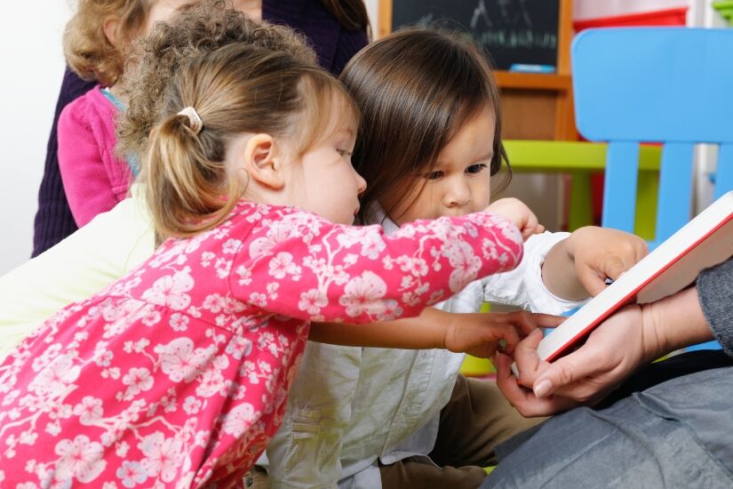 Two preschoolers point to a book held by a teacher during storytime.